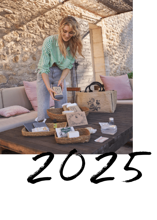 Photo d'une femme présentant des produits naturels sur une table en bois, avec des paniers et des emballages écologiques, dans un cadre extérieur ensoleillé avec un mur en pierre et une ambiance chaleureuse, ainsi que 2025 écris en lettre en dessous